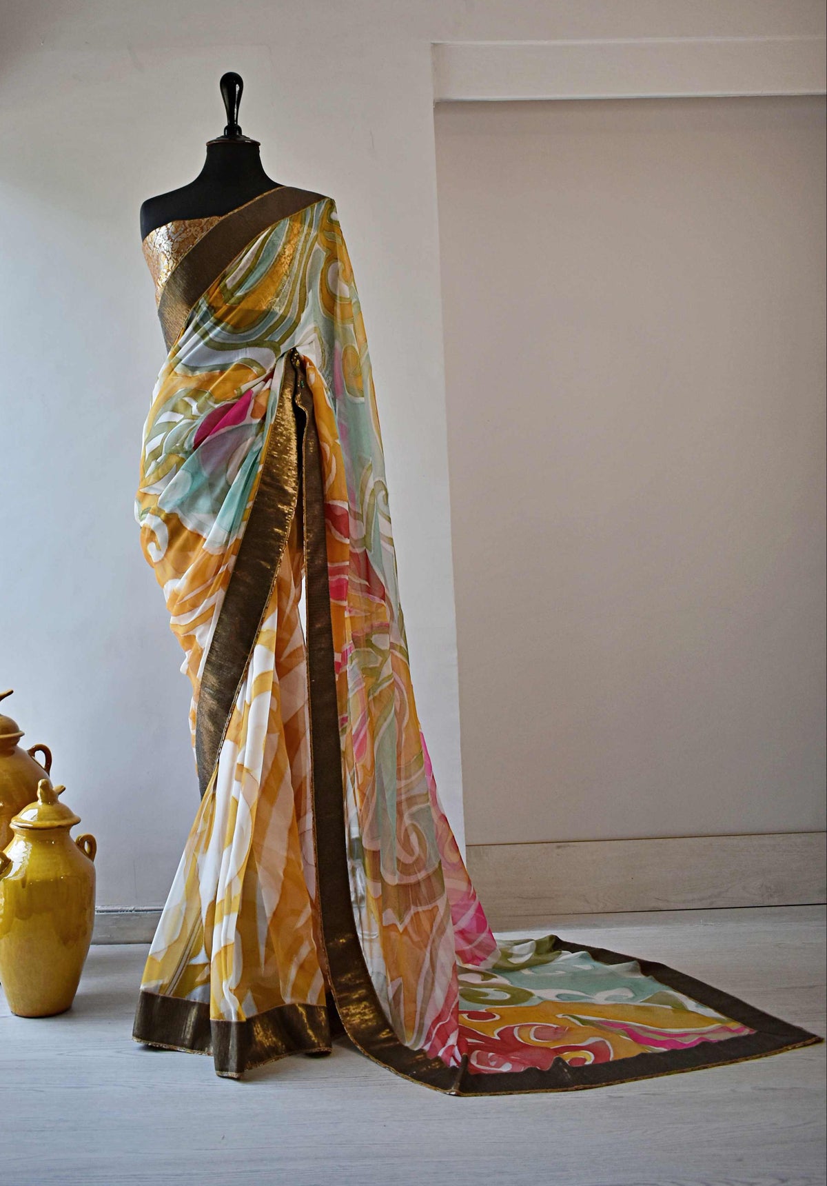 A multicolored printed saree draped on a black mannequin, featuring a yellow, pink, and green abstract pattern with a dark brown border, set against a neutral beige wall with decorative yellow ceramic vases on the floor.